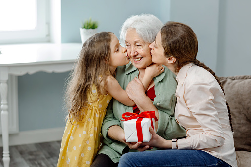 Happy Mother's Day. Daughter and granddaughter congratulate mother and grandmother, give her flowers and a gift. Grandmother, granddaughter and daughter smile and hug with a hot kiss, sitting on the sofa in the living room. Family holiday and traditions.