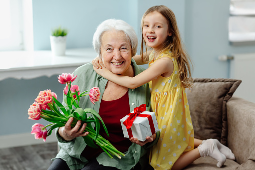 Happy granddaughter and grandmother laugh at the original congratulations on the holiday. A smiling caring granddaughter presents flowers to an elderly grandmother on women's Day.