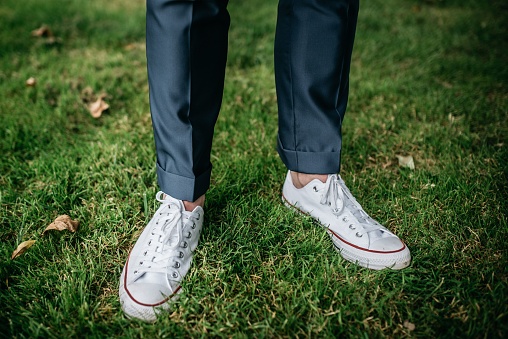 A mature man in formal attire stands in an outdoor grassy area, wearing white sneakers