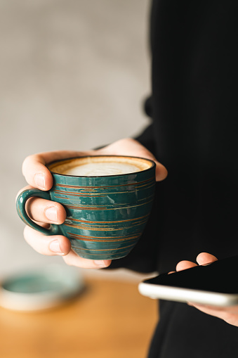 Smartphone and handmade ceramic cup of coffee in the hands of a man close-up, the concept of using technology, morning in the office.