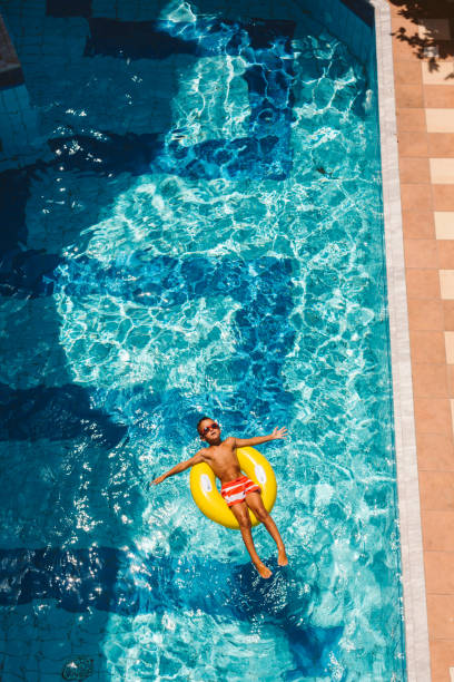 A young boy floats happily in a swimming pool on a bright yellow duck inflatable ring, wearing orange arm bands during his summer vacation trip