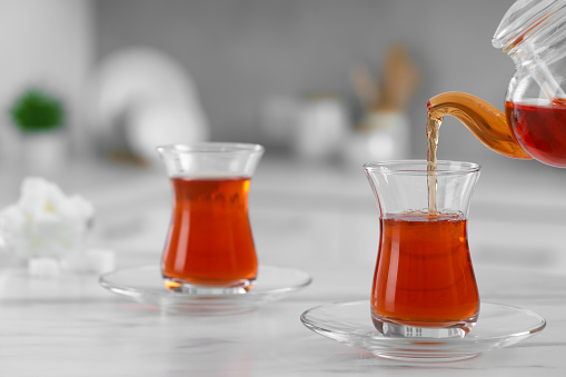 Pouring traditional Turkish tea from pot into glass on white table, space for text