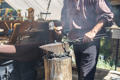 One middle-aged caucasian male blacksmith forges iron with a sledgehammer, standing behind a wooden stump on the street of Luxembourg city medieval castle, side view, close-up. The concept of medieval needlework, artisanal.