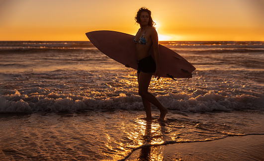 Full body female surfer in swimwear carrying surfboard and looking away while walking on beach against waving sea and sundown sky