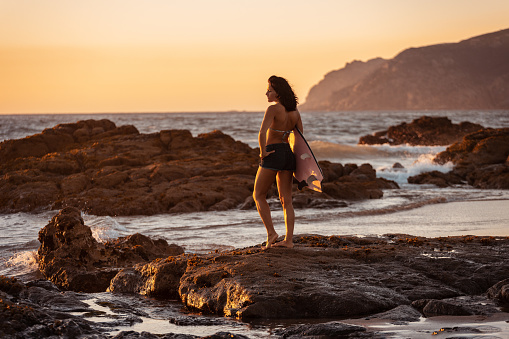 Back view of female surfer in short holding surfboard while standing on rough rock in water of ocean and looking away in sunset