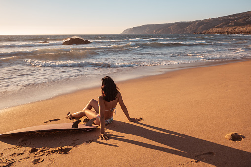 Full body unrecognizable brunette in bikini sitting on sand near surfboard and observing waving sea at sunset on beach