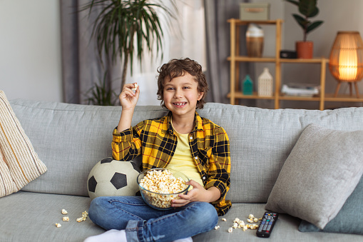 Home alone. Happy little boy having fun, watching football on tv and eating popcorn, sitting on messy sofa and laughing, free space