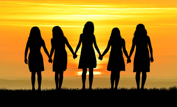 Five young women walk towards the sun at sunset holding hands