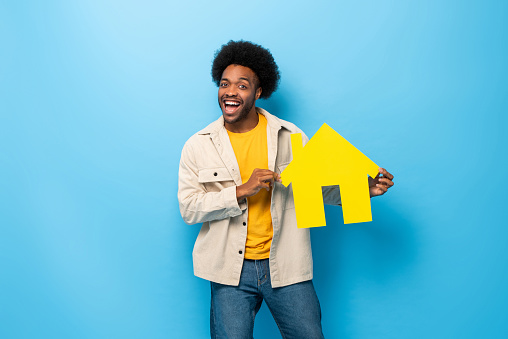 Happy smiling Afro African-American man holding home cut out in isolated light blue studio background