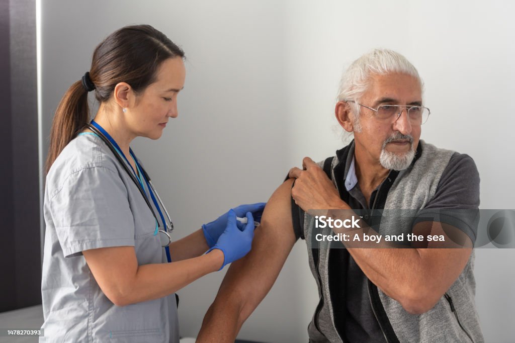 Yearly cold and flu vaccination A female healthcare worker provides a senior male patient with his yearly flu vaccine. She is wearing gloves and grey medical scrubs. The senior male of Indian ethnicity is looking in the opposite direction of the needle. Vaccination Stock Photo
