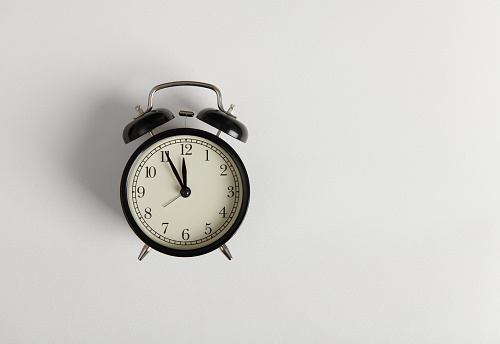 Still life composition with a black retro alarm clock showing 5 minutes to midnight or twelve o'clock on the clock face, isolated over white studio background with copy space for advertising text