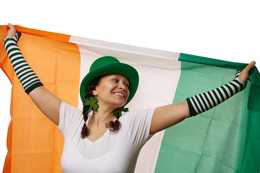 Charming Irish woman woman wearing green St Patrick's hat and clover leaves earrings, carrying Irish flag and expressing happy positive emotions, isolated over white background with free ad space