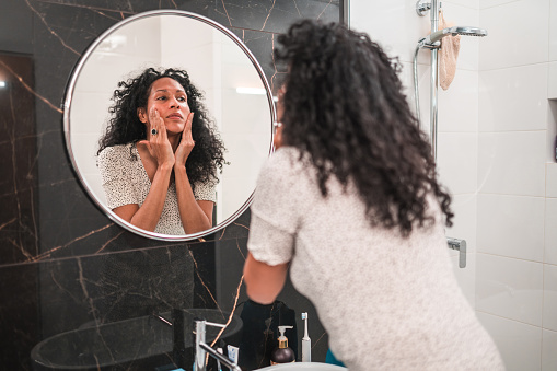 Mid adult Hispanic female standing in front of a mirror in a domestic bathrom. Taking care of her skin by applying face cream as part of a daily routine.