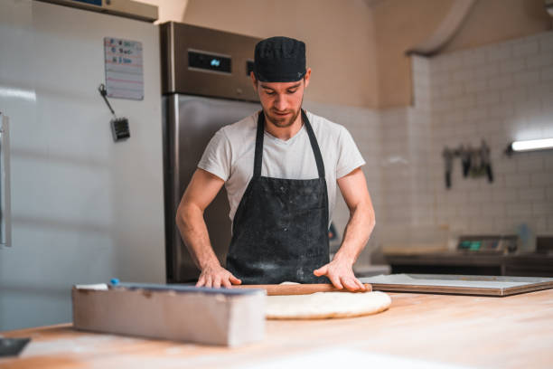 istock Caucasian Male Baker Rolling Dough On A Counter In An Artisan Baker 1478300507