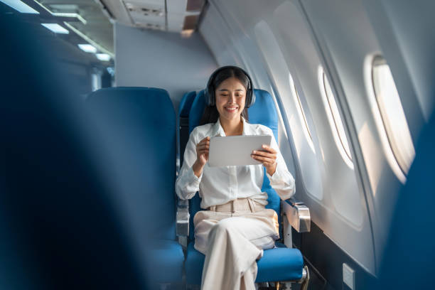 joven mujer de negocios asiática o pasajera con auriculares inalámbricos y trabajando con una tableta durante el vuelo - silla al lado de la ventana fotografías e imágenes de stock