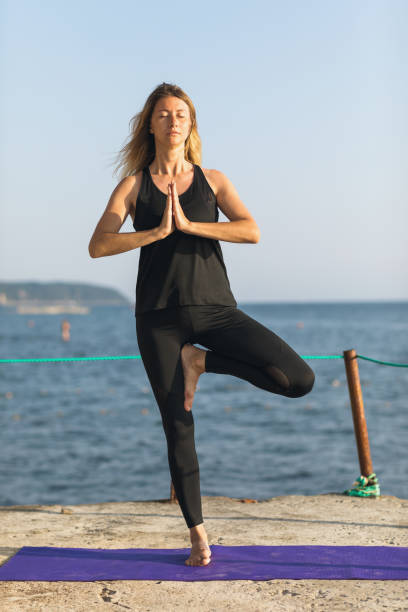 istock Woman leading a healthy lifestyle and practicing yoga, performs vrikshasana exercise, tree pose with namaste, trains in sportswear, standing on the pier by the sea on a sunny evening 1478310187