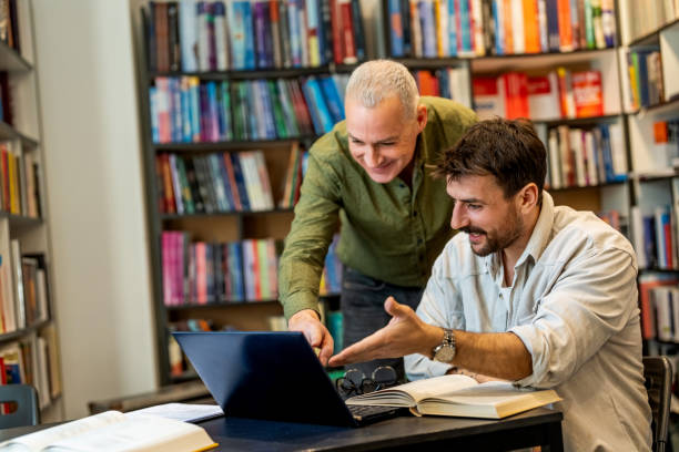 istock Adult student e-learning with help of his mature teacher in a library. 1478316099