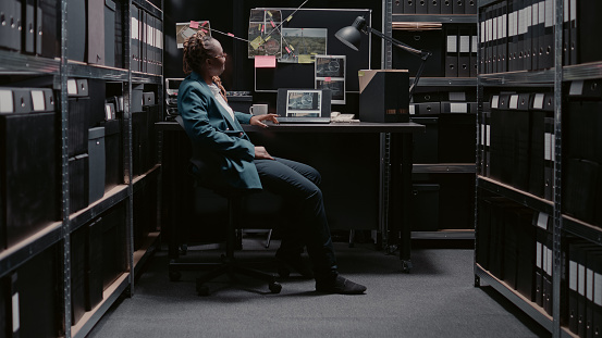African american woman examining evidence map to find suspect, using detective board on office wall. Young police officer analyzing research information for investigation, archive room.