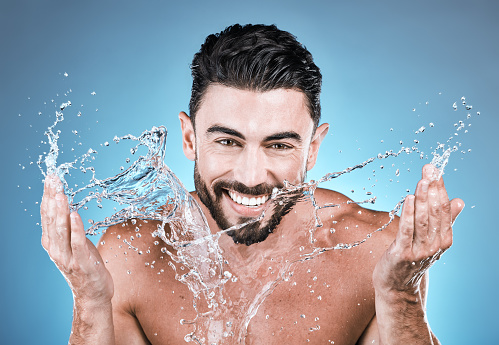Portrait, water splash and a man model washing his face in studio on a blue background for hygiene or hydration. Bathroom, skincare and cleaning with a handsome male splashing his skin for beauty