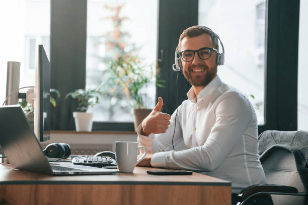 istock Smiling, sitting by table. In headphones. Office worker in formal clothes is sitting by computer and working 1478327731