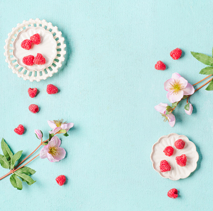 Food background with fresh raspberries in white bowls on light turquoise background with flowers,  top view with copy space