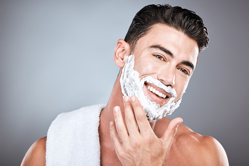 Applying, shaving and portrait of a man with cream on face isolated on a grey studio background. Happy, hygiene and headshot of a person with a facial product for beard hair removal on a backdrop