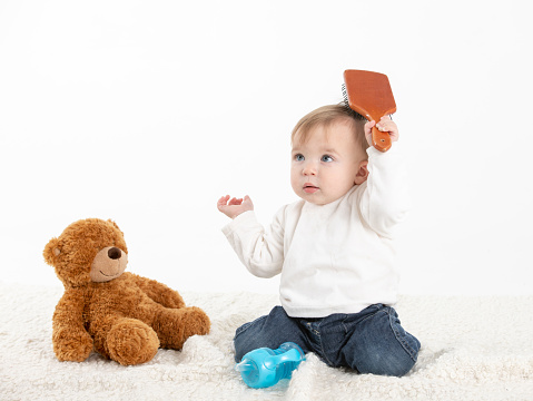 Stock studio photo with a white background of a baby combing his hair with a teddy bear by his side.