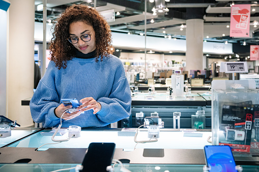 Positive young African American female in casual outfit and glasses using and selecting smartphone in modern electronics store