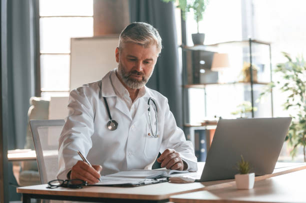 istock Sitting by table with laptop, writing data into document. Senior doctor in white coat is at work indoors 1478344560