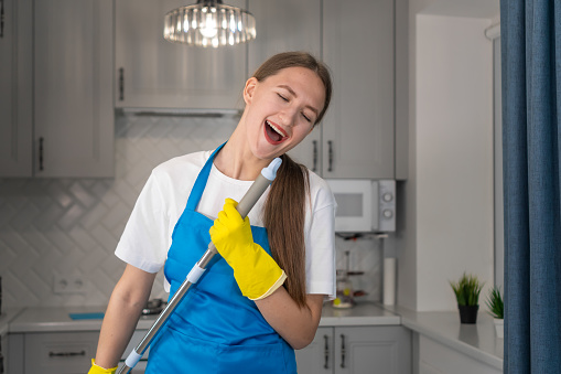 Beautiful Caucasian female comedian in uniform in an apron, white T-shirt dancing, singing with a mop while cleaning the kitchen in the house. Happy professional cleaning company girl enjoys life