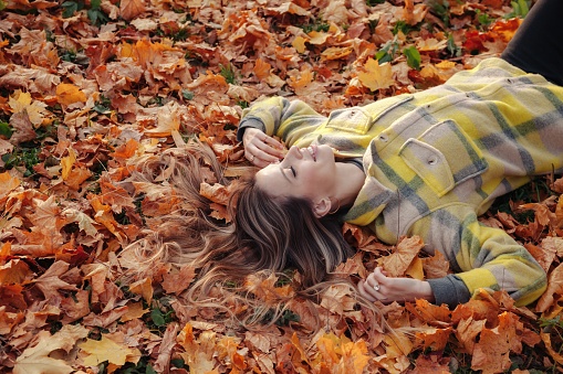 Cute young woman lying down on fall leaves in autumn park spend time outdoors, smiling happy. Pretty lady in yellow coat relaxing in nature. Concept of relax and leisure activity. Copy ad text space