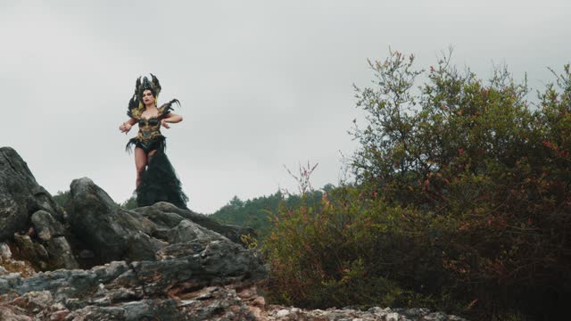 a witch in a black gown and crown dances on a rock in a mountain
