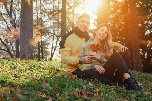 Lovely couple sitting on grass in autumn park, sunshine outdoors, smiling happy. Cute couple, man and woman, in yellow clothes rest in nature. Concept of relax and leisure activity. Copy ad text space