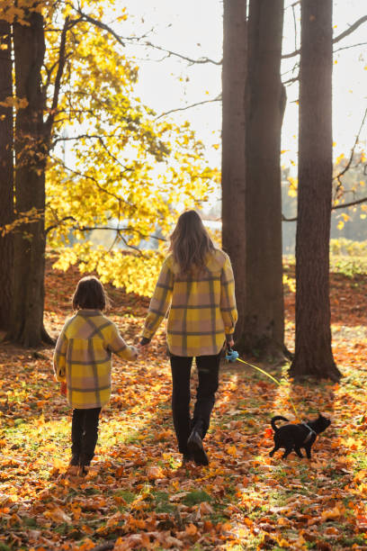 Rear view of mom and daughter with doggy in yellow coats walk together in autumn park outdoors, family fun. Cute kid with mother posing in fall nature. Concept of leisure activity. Copy ad text space vista trasera de mamá e hija con perrito en abrigos amarillos caminan juntas en el parque de otoño al aire libre, diversión familiar. lindo niño con madre posando en la naturaleza otoñal. concepto de actividad de ocio. copiar el espacio de texto del a