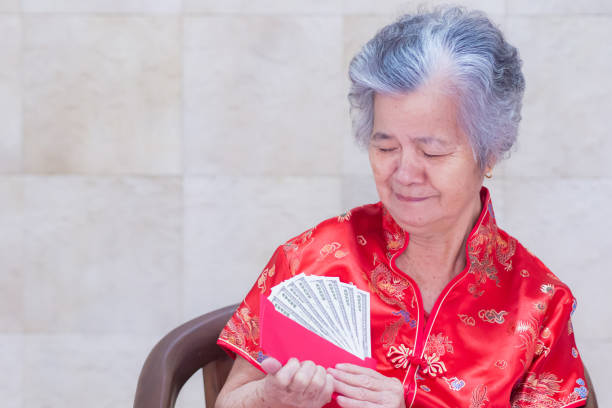 anziana donna asiatica che indossa una camicia rossa cheongsam con in mano un pacchetto di buste rosse con dollari usa. - cheongsam chinese culture indoors looking at camera foto e immagini stock