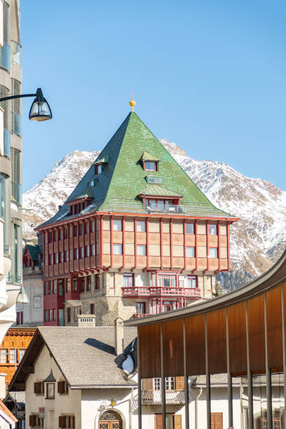 View of alpine residential houses built on a snowy hillside, surrounded by evergreen trees, with a Swiss national flag visible on one of the buildings during winter daylight.