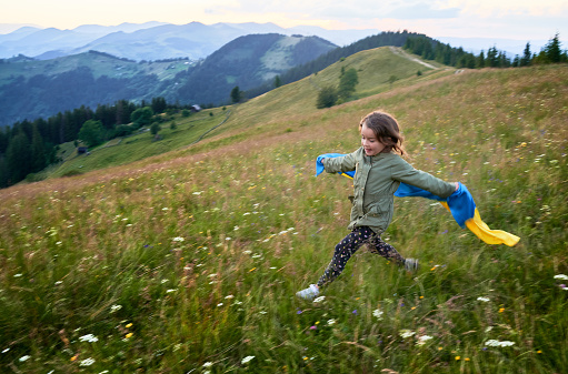 Cute girl running in mountains with Ukrainian flag. Little girl smiling at camera with Ukrainian flag on her shoulders. Feeling freedom in young people.