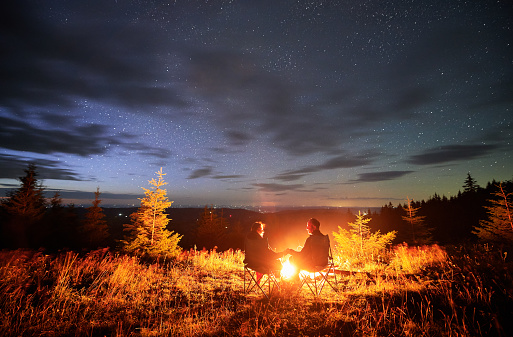 Happy couple staring at starry sky. Young people sitting in chairs and warming by campfire. Travelling couple having a rest together. Night camping.