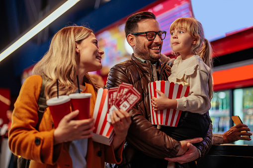 A young  Caucasian family is in the cinema with their small child, carrying soda, popcorn and tickets on their way to the movie hall.