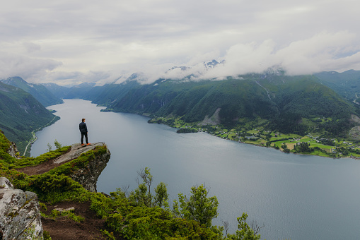 Male hiker getting to the edge of the cliff contemplating dramatic view of sea and green hills of the beautiful fjord in Western Norway