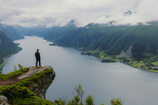 Male hiker getting to the edge of the cliff contemplating dramatic view of sea and green hills of the beautiful fjord in Western Norway