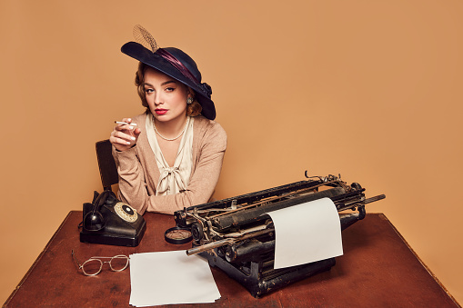Calmness. Attractive woman writer wearing old-fashioned clothes sitting at table and smoking over beige background. Concept poems, novel, emotions, beauty, fashion, retro style, vintage