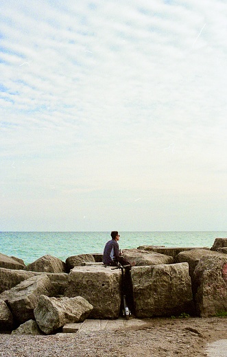 Toronto, Canada – December 16, 2021: A guy sitting on coastal rocks overlooking the horizon at Kew Balmy Beach, Toronto, Canada