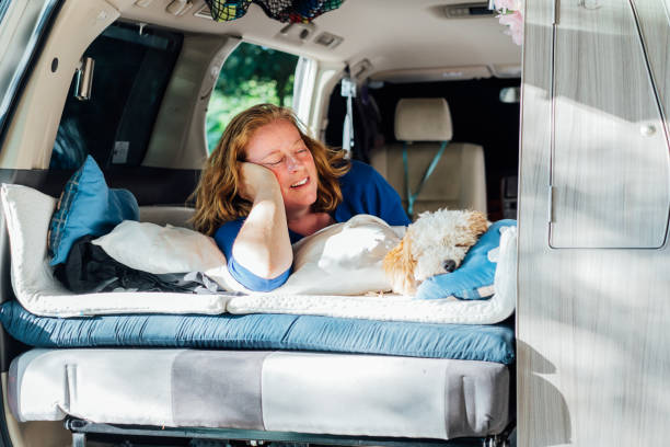 istock Smiling middle-aged woman looking on sleeping cockapoo puppy pet while lying in camper car vehicle during road trip. Window view. Best friend. Enjoying free lifestyle, travel, vacation, freedom. 1478404072