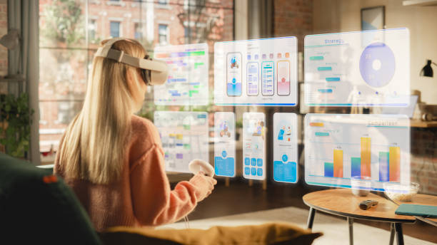 istock Female with Blond Hair Using Virtual Reality Headset and Controllers at Home. Creative Woman Sitting on a Couch in Living Room Working on a Task and Checking Charts, Statistics and Schedule 1478408512