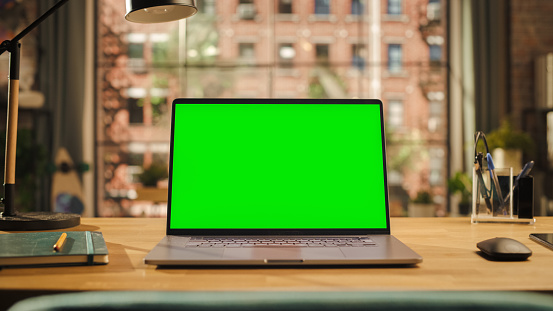 Laptop Computer Display with Mock Up Green Screen Standing on a Wooden Desk in Living Room. Chroma Key Monitor at Home or Creative Loft Office With Notepad, Mouse and Pencil