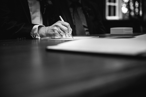 A mature Caucasian male is sitting at a desk in a professional setting, signing a document