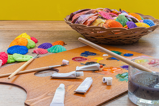 A closeup of a variety of paints and brushes on a wooden palette with seashells on the table