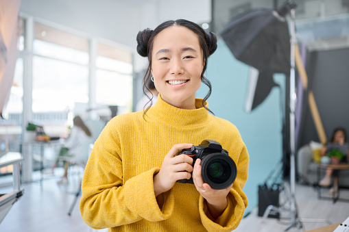 Smile, camera and portrait of a photographer working at a studio for a photoshoot. Happy, media and creative Asian woman shooting, filming or learning about photography production as a career