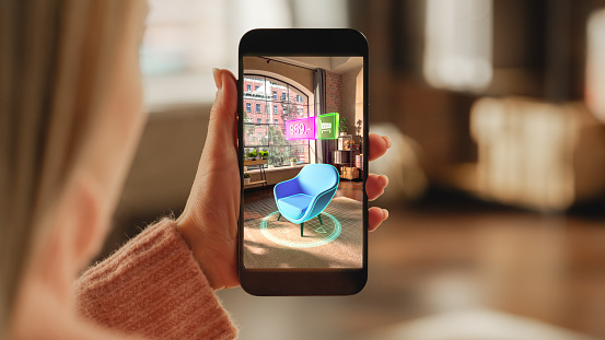 Over the Shoulder Footage of a Female Hand, Holding Smartphone with an Augmented Reality Display Showing a Chair. Woman Doing Online Shopping and Checking her Options In Live Situation In Distance